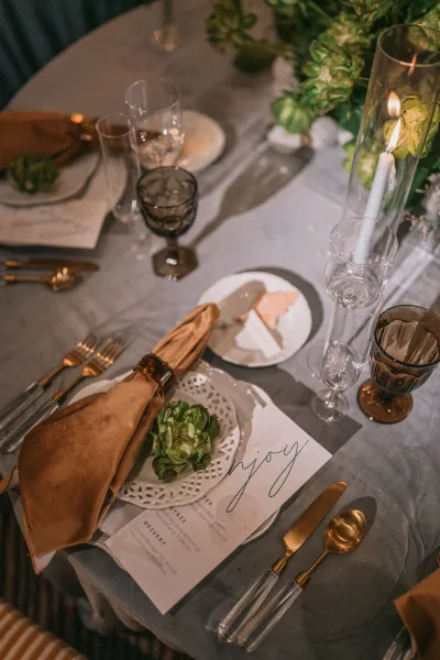 Reception tablescape with wedding place setting, greenery centerpiece and taper candles in a glass hurricane on a gray tablecloth with mixed glassware