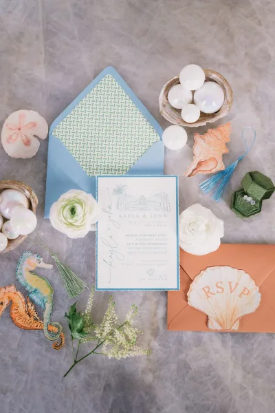 Invitation suite with beach wedding invitations, envelopes, RSVP card and tassels beside a velvet ring box, pearls and seashell dish on gray fabric backdrop