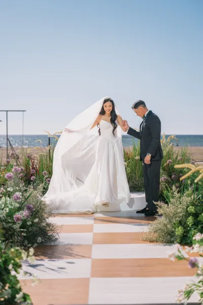 Couple portrait of bride and groom holding hands, her veil blowing on an oceanfront aisle with greenery and purple flowers under blue sky
