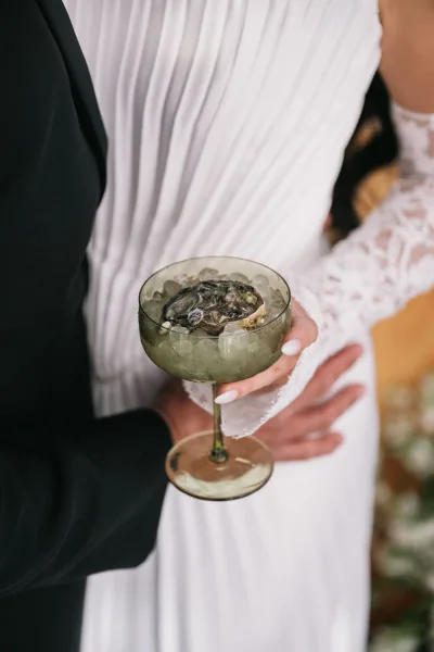 Wedding cocktail oyster martini in a coupe glass with ice and an oyster garnish, held by bride in lace sleeve beside groom in black suit indoors