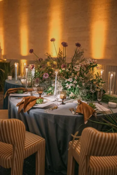 Reception tablescape with wedding table centerpiece on a blue tablecloth, taper candles in glass hurricanes, greenery garland, warm uplighting