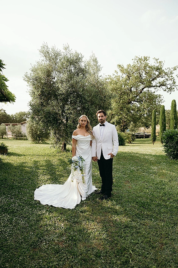 Couple portrait of bride and groom holding hands, her off-shoulder gown and bouquet beside his white dinner jacket on a lawn with olive trees