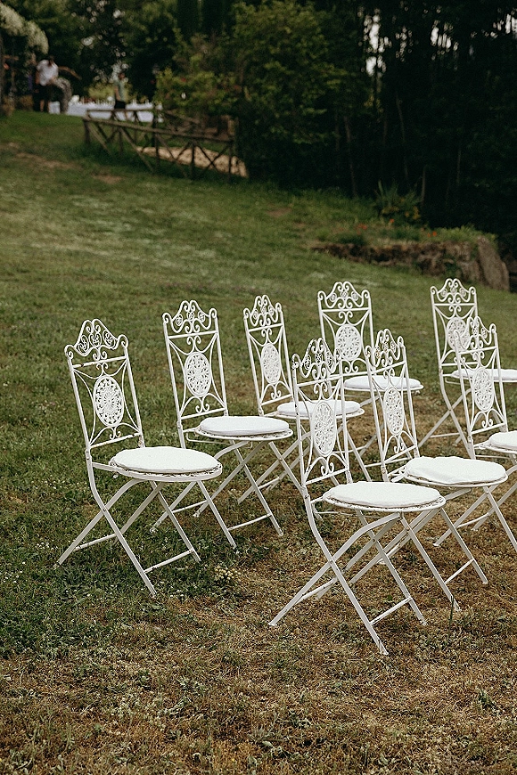 Ceremony seating with outdoor ceremony chairs, white metal chairs and cushions arranged on a grassy lawn beside trees and a wooden fence