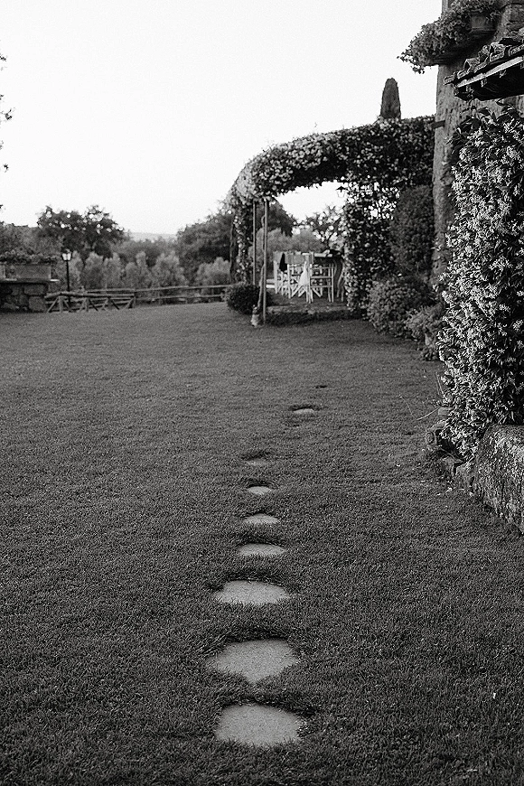 Outdoor ceremony setup with garden wedding ceremony chairs lining a stepping-stone aisle to a floral arch on a lawn, framed by hedges and hills