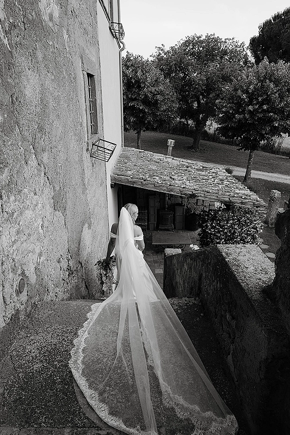Bridal portrait of a back view bride in a cathedral veil, holding a bouquet and descending stone steps in a rustic courtyard setting