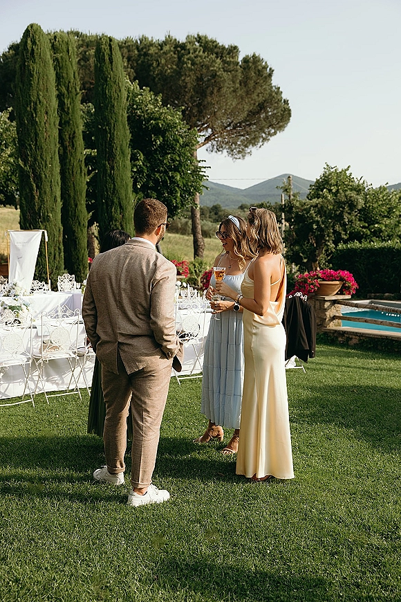 Wedding cocktail hour with guests mingling by a pool, holding champagne coupe glasses on a garden lawn with cypress trees and mountain views
