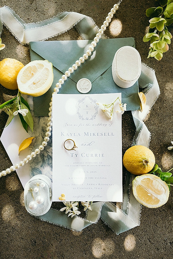 Wedding invitation suite flat lay with dusty blue cards, sage envelope, wax seal, silk ribbon, pearls and rings on sunlit stone surface