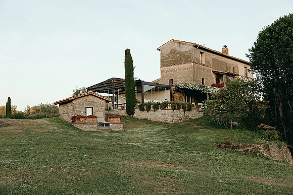 Wedding venue exterior with rustic wedding venue stone buildings, pergola and vine-covered terrace, outdoor chairs on a grassy hillside under open sky