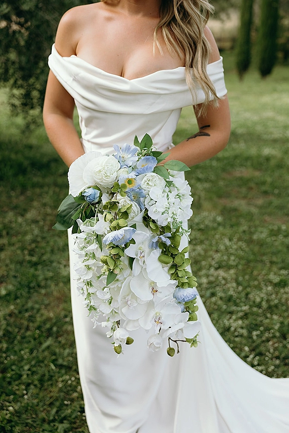 Bridal bouquet with cascading orchid bouquet of white orchids, roses, blue flowers, and greenery held against an off-shoulder gown on a garden lawn