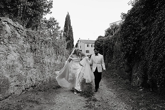 Couple portrait of bride and groom walking, she lifts her dress train as her veil blows along a gravel path by a stone wall