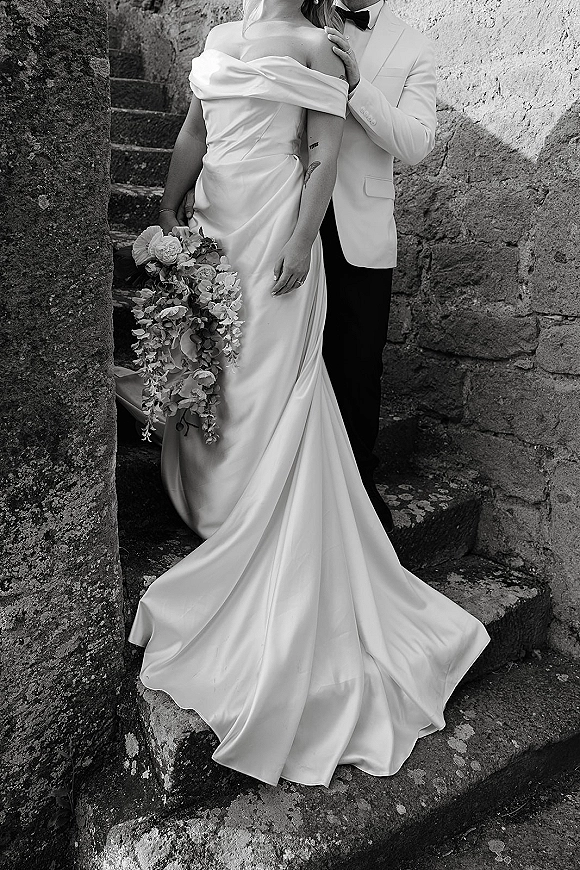 Couple portrait in a black and white wedding portrait as groom embraces bride in satin off-shoulder gown with bouquet on stone stairs