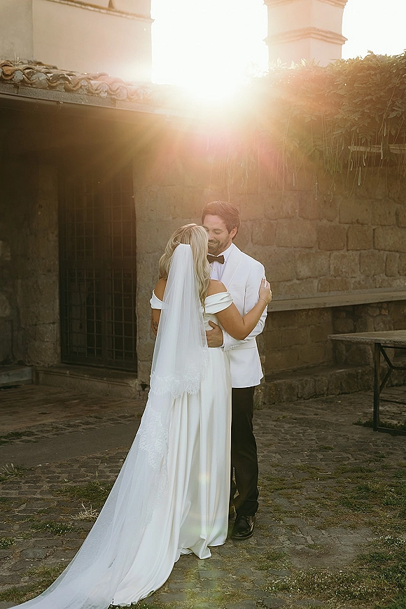 Wedding couple portrait of bride and groom embrace, her cathedral veil glowing in sun flare beside an ivy stone wall in a cobblestone courtyard