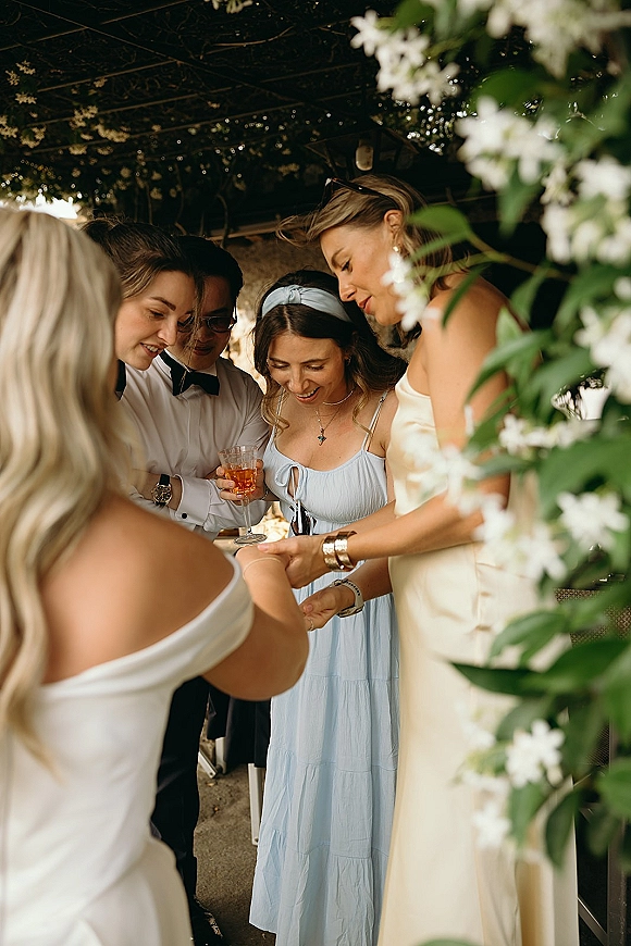 Wedding guest candid of bridesmaids mingling with a cocktail glass and bow tie under shady foliage along a walkway with white flowers