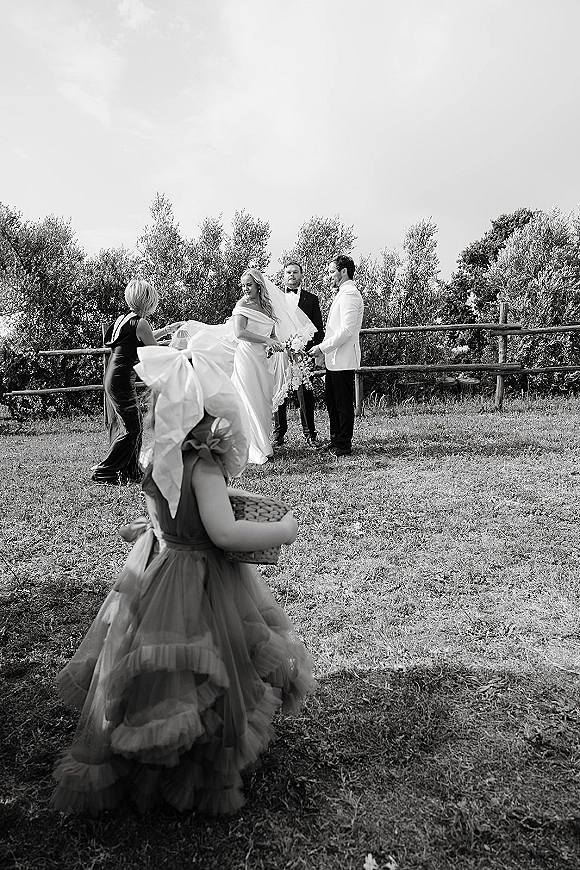 Ceremony moment at an outdoor wedding ceremony with bride and groom at the altar, veil blowing as flower girl holds a basket on lawn by rustic fence