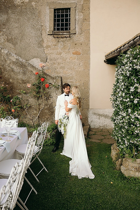Couple portrait of bride and groom outdoors, she holds a cascading bouquet in a courtyard by stone wall and reception table with roses