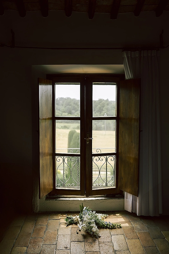 Bridal bouquet of white and blue bouquet blooms with greenery, resting by an open wooden window with wrought iron balcony railing