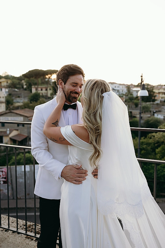 Couple portrait of bride and groom embrace on a balcony, her long veil and satin dress flowing as he smiles in a tuxedo above hills.