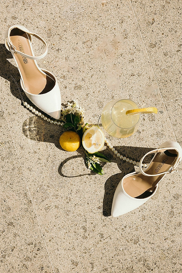 Wedding shoes flatlay with white ankle strap heels, pearls, engagement ring and lemon cocktail on sunlit stone pavement with shadows
