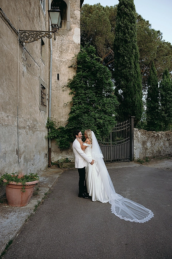 Wedding couple portrait of bride and groom embrace by an ivy-covered stone wall, her long veil train flowing near a wrought iron gate