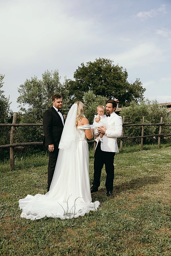 Ceremony moment at an outdoor wedding ceremony with bride in off-shoulder gown and veil beside groom holding baby by a wooden fence