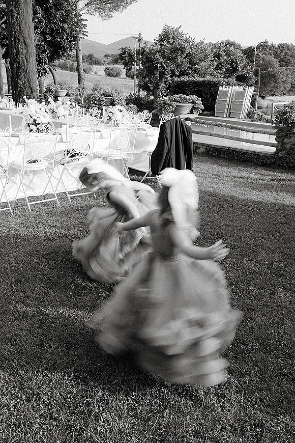 Wedding reception kids running and twirling between round tables with floral centerpieces on a lawn, with trees and stone wall behind