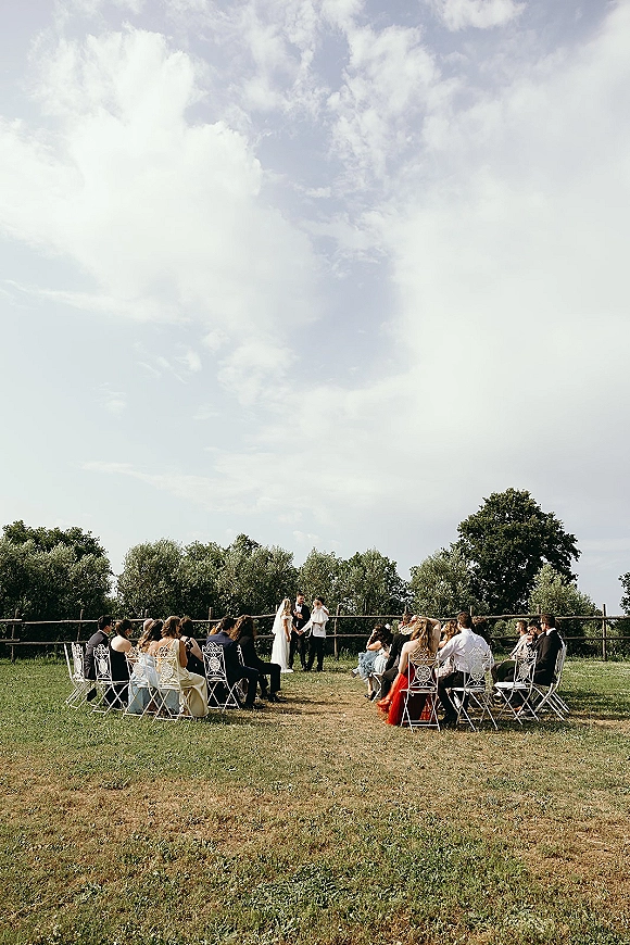 Wedding ceremony with bride and groom facing an officiant, guests seated on white chairs in a semicircle on a grassy lawn by trees and fence