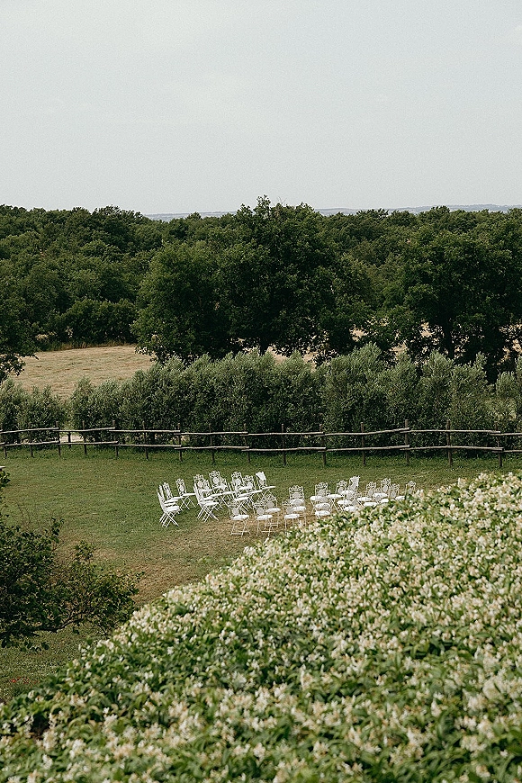 Ceremony setup with white folding chairs arranged in a semicircle on a grassy lawn, framed by a wooden fence, trees, and rolling hills