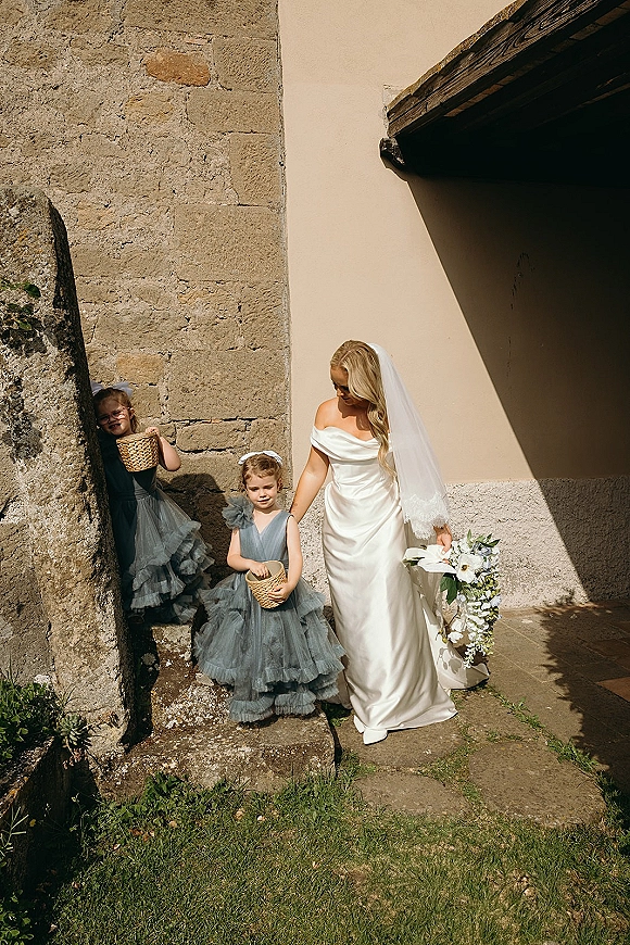 Bride with flower girls holding flower girl baskets, carrying a cascading white bouquet and veil by stone steps in sunlit shadows