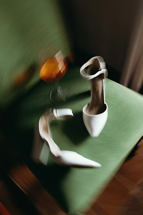 Bridal shoes, white wedding heels with ankle straps resting on a green chair over a warm wood floor, pointed toes neatly aligned