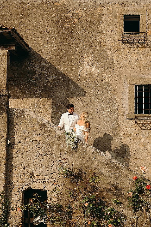 Couple portrait of bride and groom looking at each other, bride holding cascading rose bouquet on stairs by a stone villa balcony