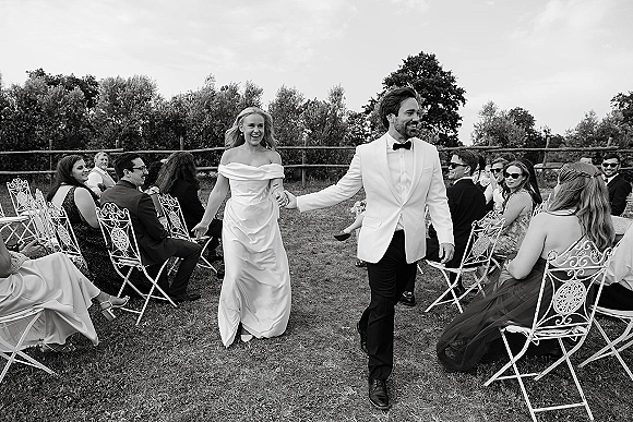 Wedding recessional as bride and groom walk the aisle holding hands between ceremony chairs on a grassy lawn, guests in sunglasses cheering under trees