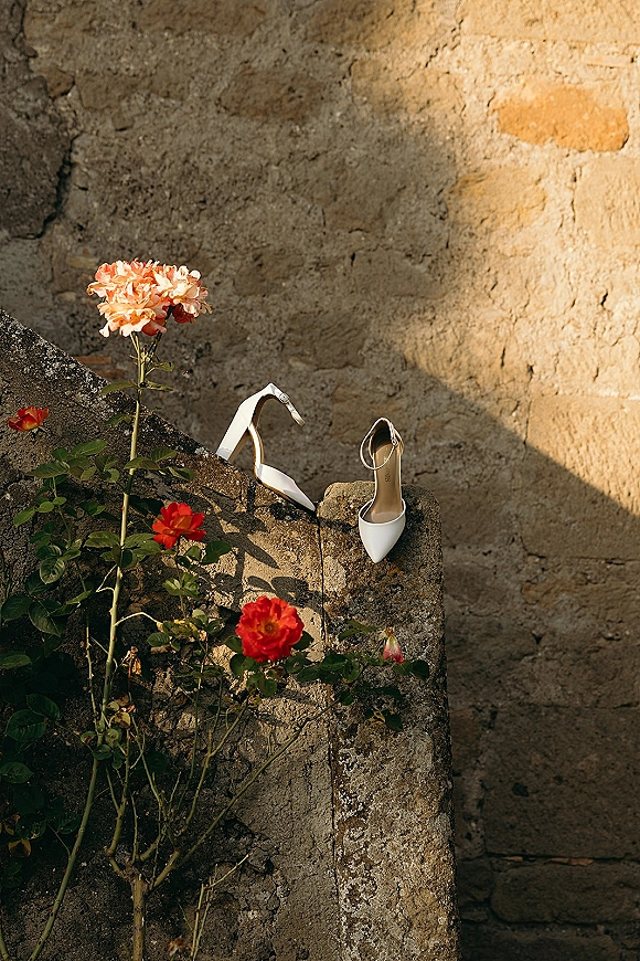 Wedding shoes with roses, white bridal heels on a sunlit stone ledge against a stone wall with greenery and shadows
