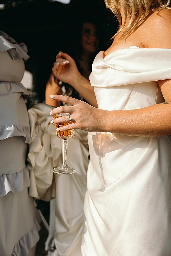 Bride portrait in an off the shoulder wedding dress holding a champagne coupe, showing engagement ring and manicure among guests in shadows