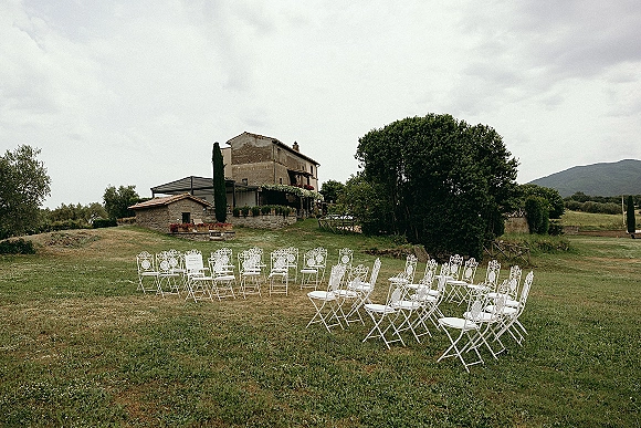 Ceremony setup with white metal chairs in a semicircle on a grass lawn, backed by a stone farmhouse and rolling hills under clouds