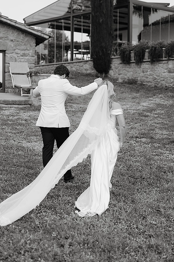 Newlywed exit as bride and groom walk away holding hands, her long lace-trim veil blowing behind, past a stone wall by a glass venue