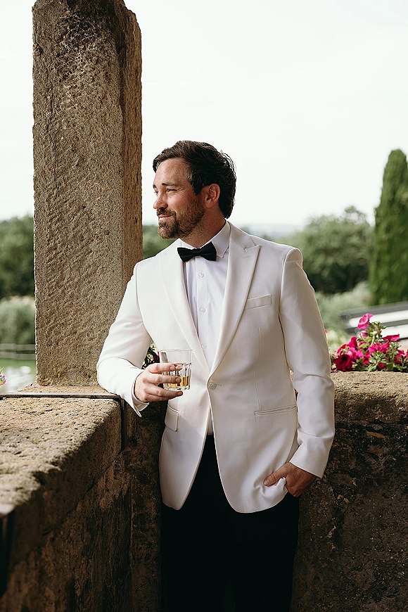 Groom portrait in a white tuxedo groom look, wearing a black bow tie and boutonniere, holding a whiskey glass on a stone terrace with greenery.