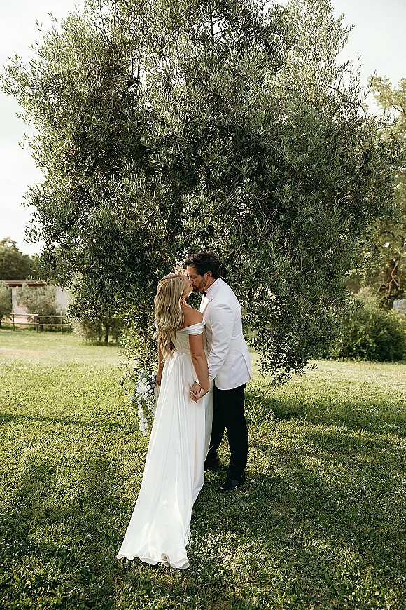 Wedding kiss portrait of bride and groom kissing, holding hands under an olive tree on a grassy lawn, bouquet in hand and white tux jacket