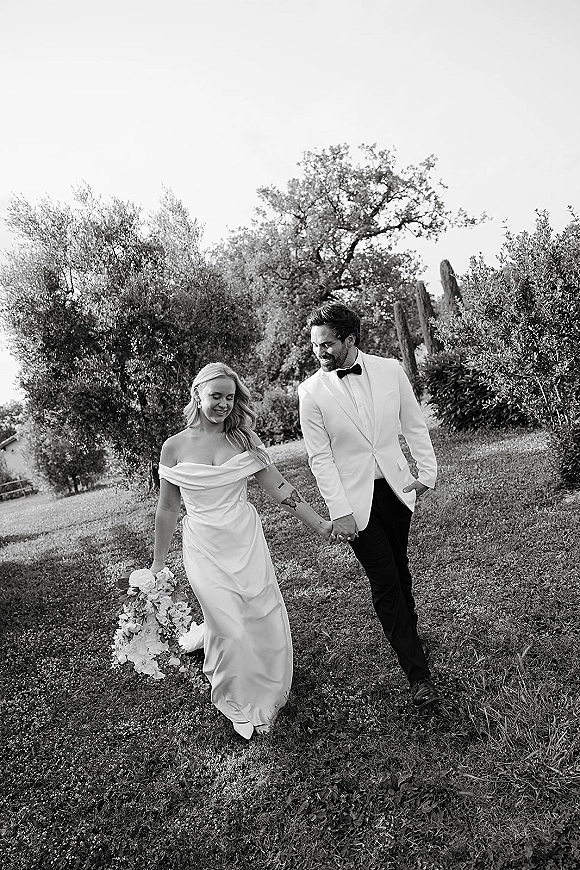 Couple portrait in a black and white wedding portrait, newlyweds holding hands and smiling on a lawn with trees, bouquet accent