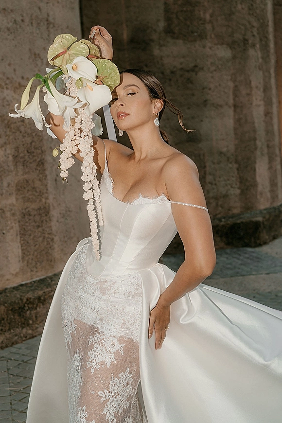 Bridal portrait of bride holding bouquet of calla lilies and anthurium, in off shoulder lace dress by a stone wall outdoors