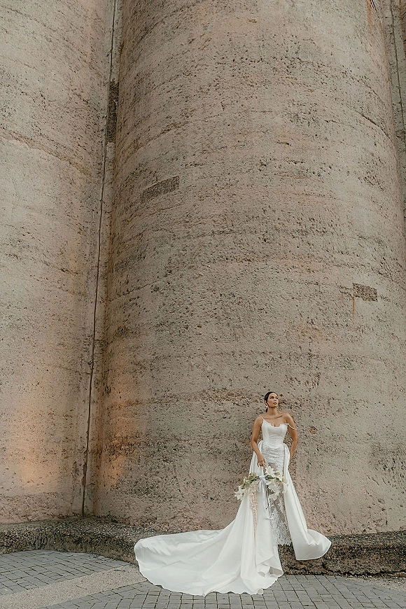 Bridal portrait of a bride in a strapless wedding dress with a long train, holding a bouquet with ribbon against an industrial concrete wall