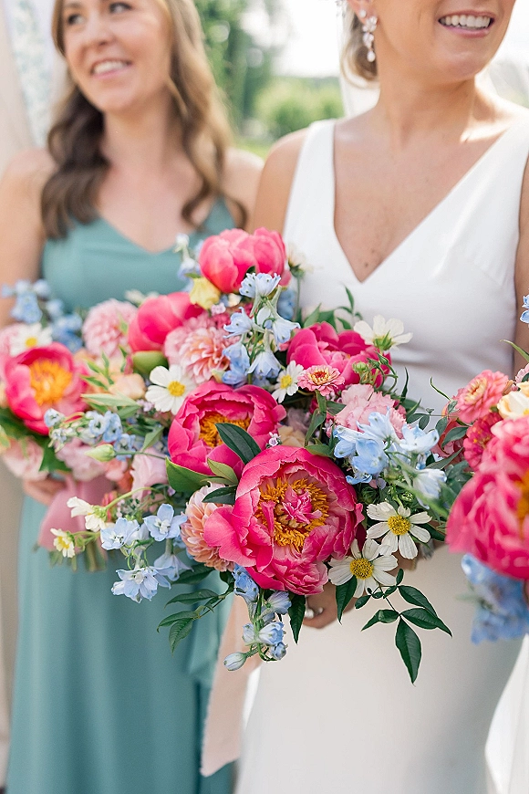 Bridal bouquet with pink peony bouquet blooms, blue delphinium, daisies and greenery, held by bride in white dress outdoors in daylight