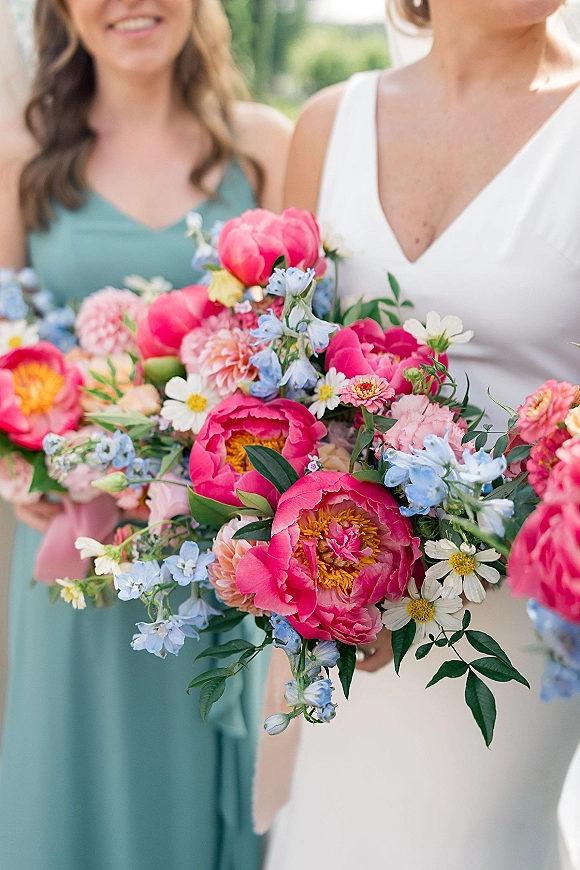 Bridal bouquet with pink peony bouquet blooms, blue delphinium, and white daisies tied with a pink ribbon against blurred greenery outdoors