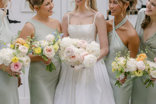 Bride with bridesmaids laughing in sage green dresses, holding garden-style bouquets beside a white wall and doorway in natural light