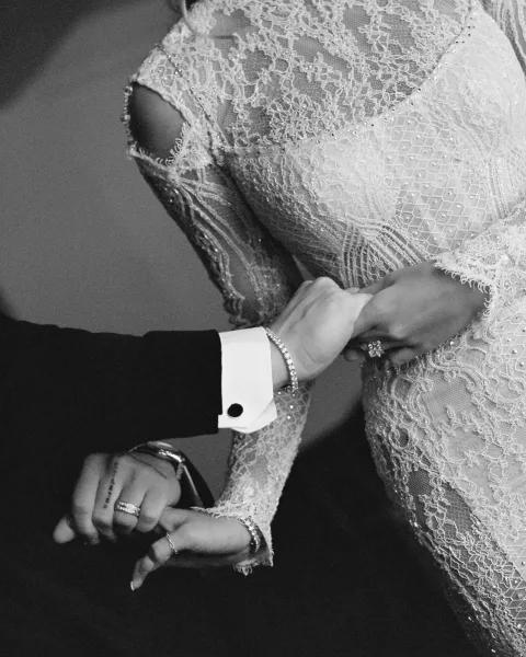 Wedding dress close-up shows lace wedding dress detail with illusion neckline and beaded bodice as couple holds hands, rings visible against wall