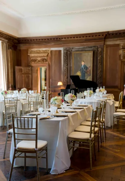 Reception tablescape with white linens, gold chiavari chairs, floral centerpieces and taper candles in a wood-paneled room with grand piano
