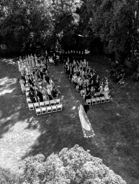 Wedding processional as bride walking down aisle in a long cathedral veil past white folding chairs on a garden lawn with trees and brick wall