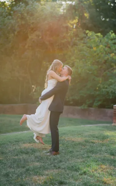Wedding kiss portrait of groom lifting the bride for a kiss, strapless dress and ring catching sunlight on a garden lawn by a stone wall