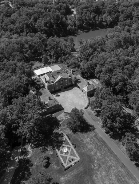 Wedding venue aerial view of a manor house with white event tent, ceremony chairs and fountain courtyard beside lawn, pond and trees