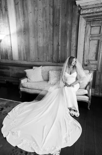 Bridal portrait of a bride on sofa in a strapless gown with long lace train and veil over face, set against wood paneling and ornate door