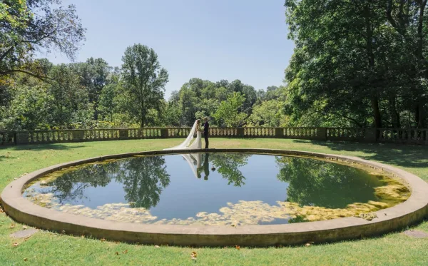 Couple portrait of bride and groom by pond, her long wedding veil train trailing as they stand on a stone edge under blue sky